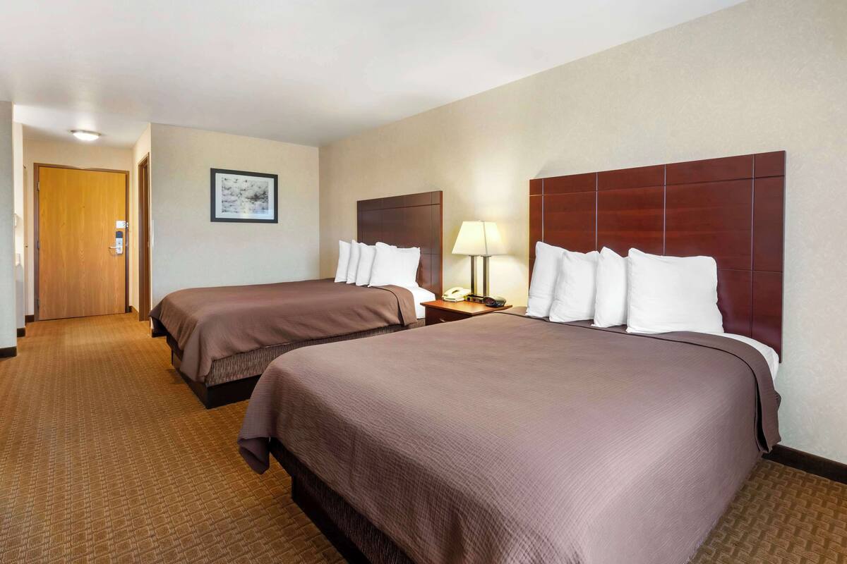 A hotel room with two beds, a wooden door, a framed picture on the wall, and a nightstand with a lamp.
