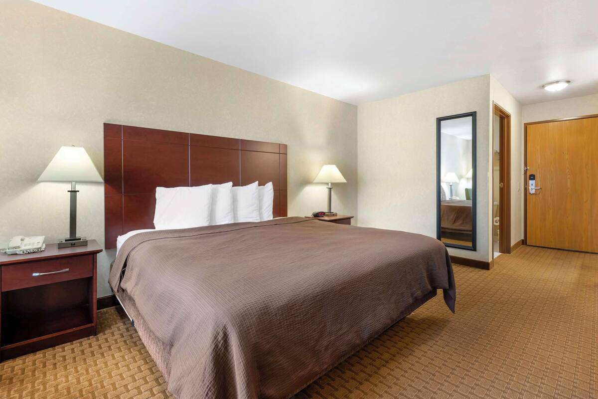 A hotel room with a large bed, two bedside tables with lamps, a wooden headboard, a mirror, and a door.