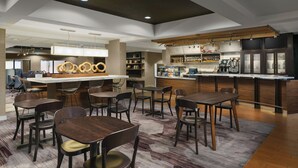 A modern restaurant with a bar area, wooden tables and chairs, and a patterned carpet.