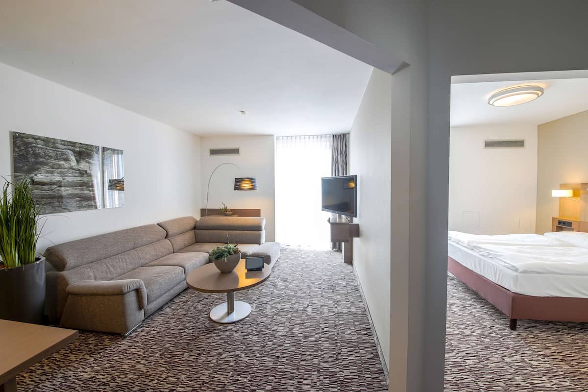 Ein Hotelzimmer mit einer Couch, einem kleinen Couchtisch, einem Fernseher, einem Bett und einem großen Fenster.