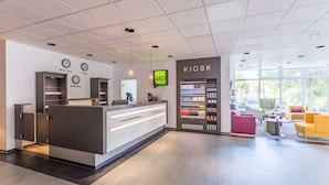 Un área de recepción moderna con mostrador, reloj digital, quiosco y zona de estar con sillas coloridas.
