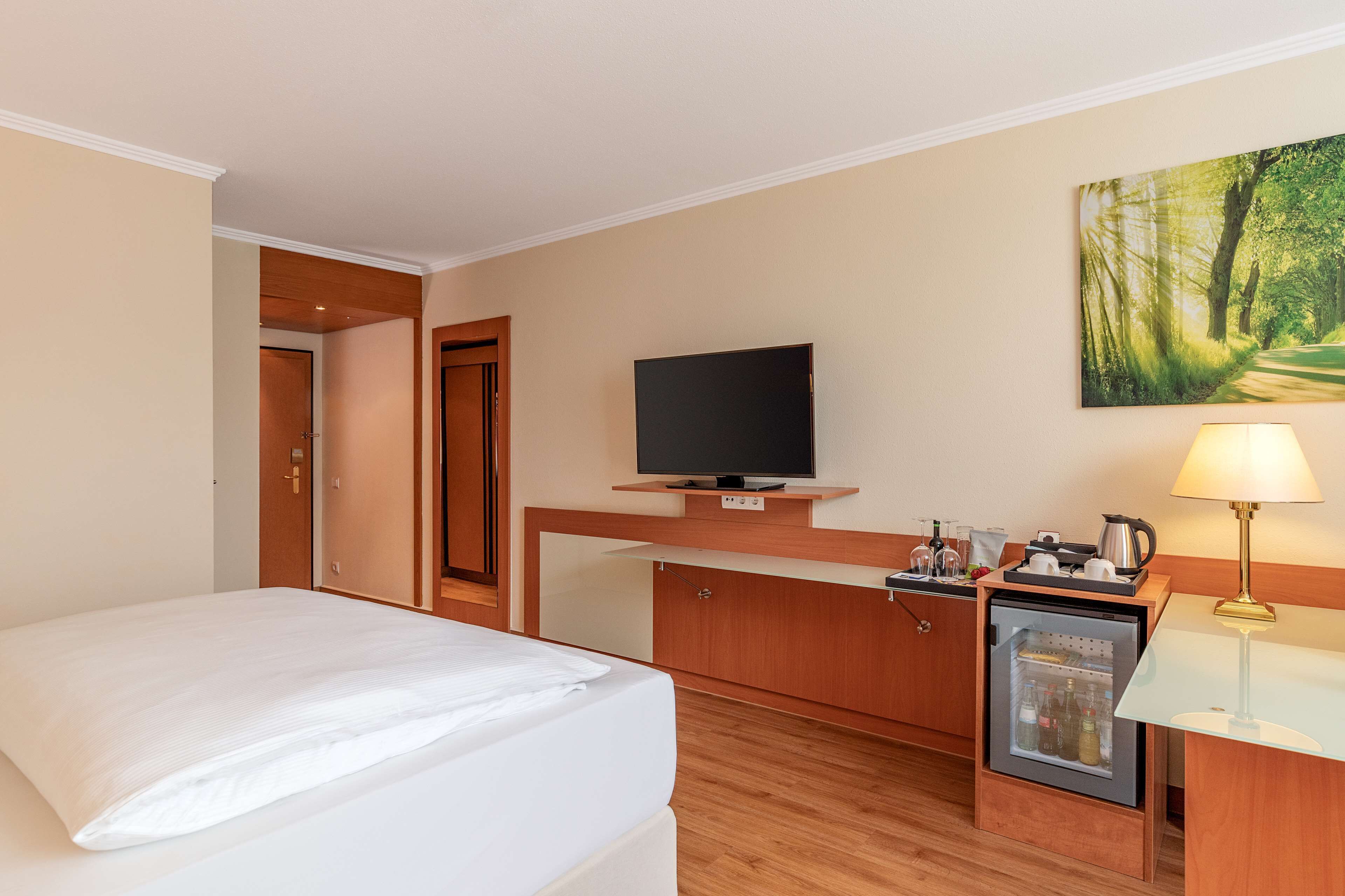 Habitación de hotel con una cama, televisión, refrigerador pequeño, escritorio con lámpara y un cuadro en la pared.