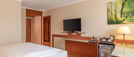 Habitación de hotel con una cama, televisión, refrigerador pequeño, escritorio con lámpara y un cuadro en la pared.