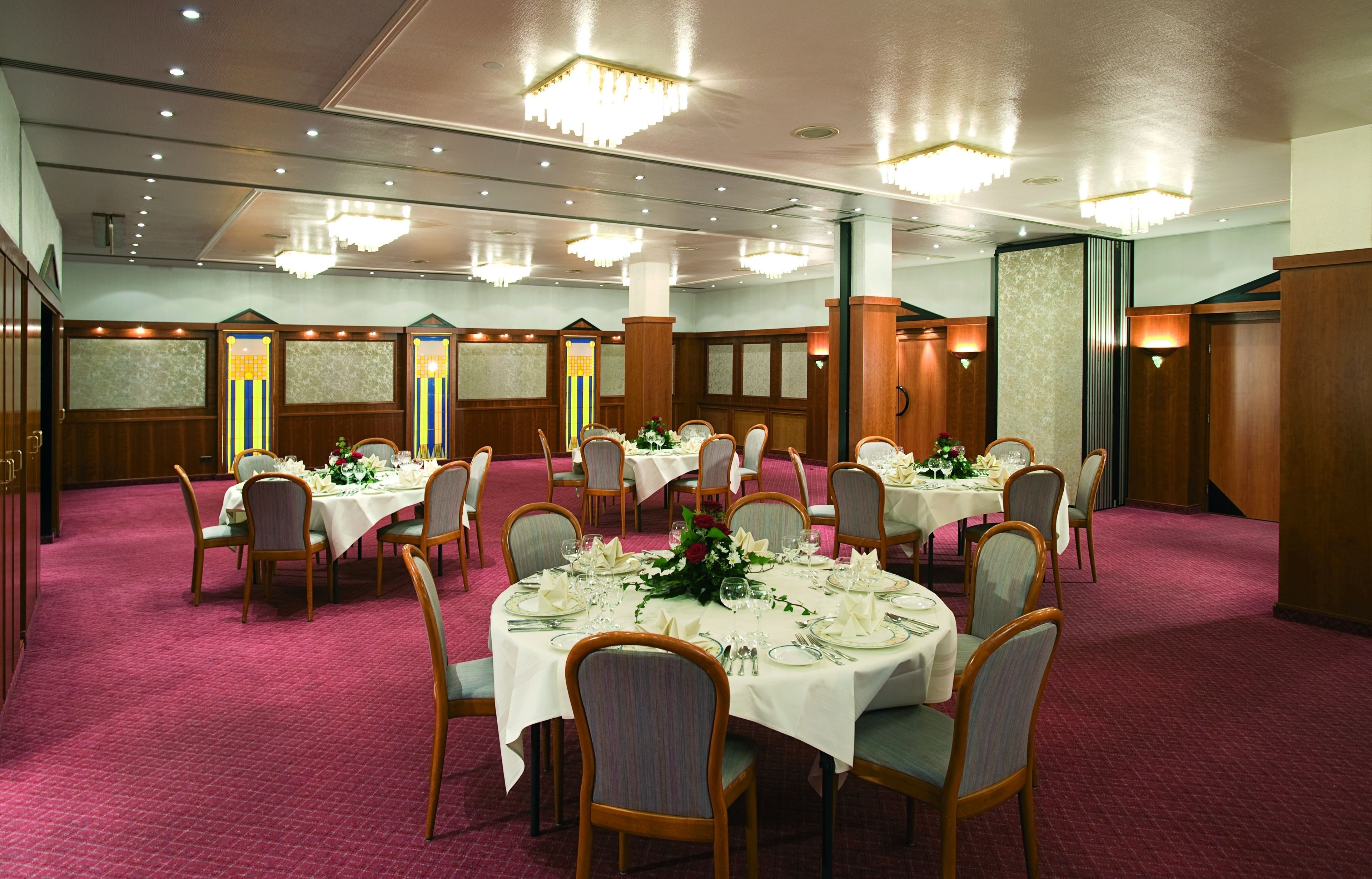 Salão de banquetes com mesas redondas preparadas para refeições, painéis de madeira e lustres.