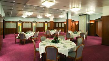 Salão de banquetes com mesas redondas preparadas para refeições, painéis de madeira e lustres.