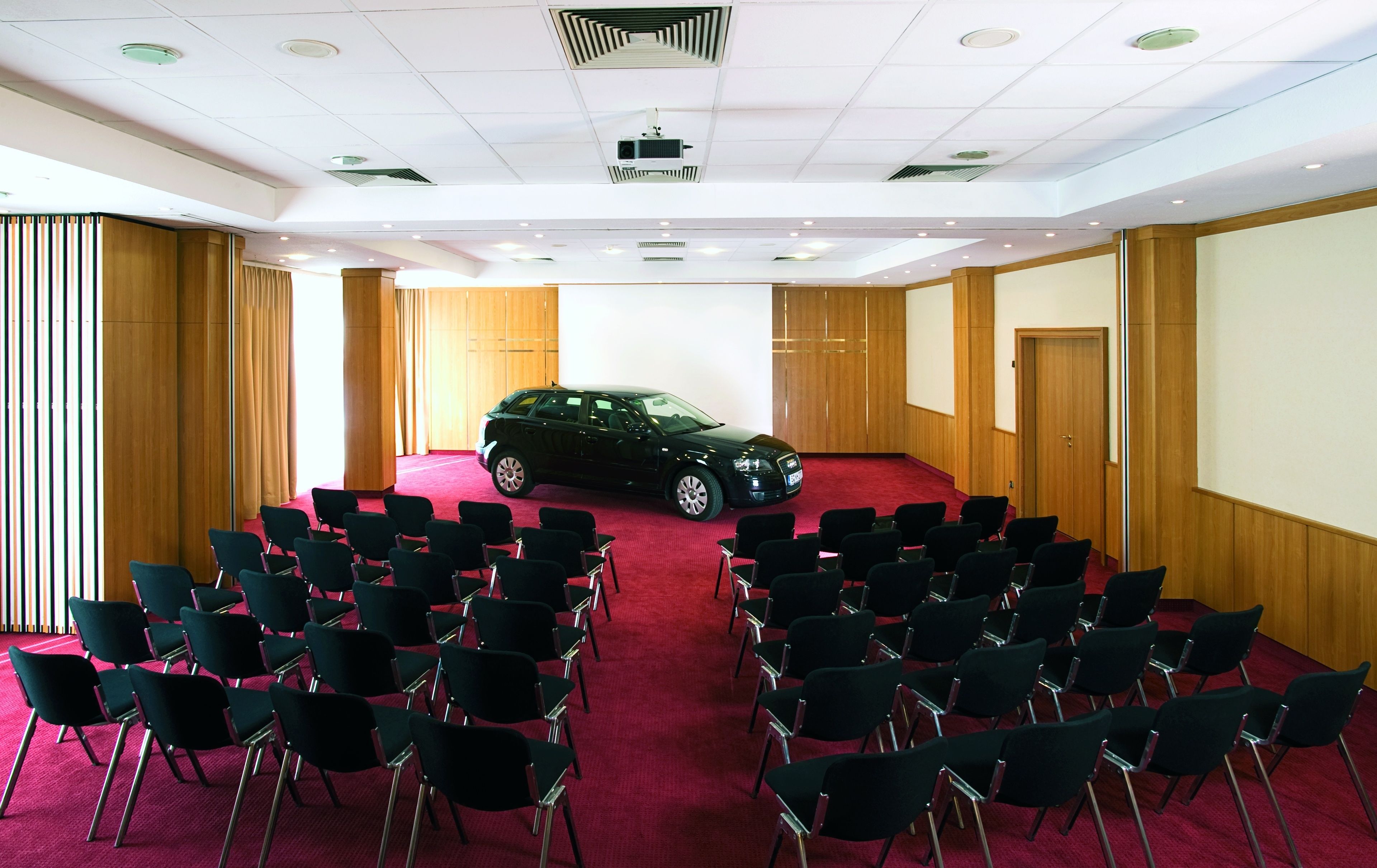 A conference room with rows of chairs, a projector screen, and a car parked in the center.