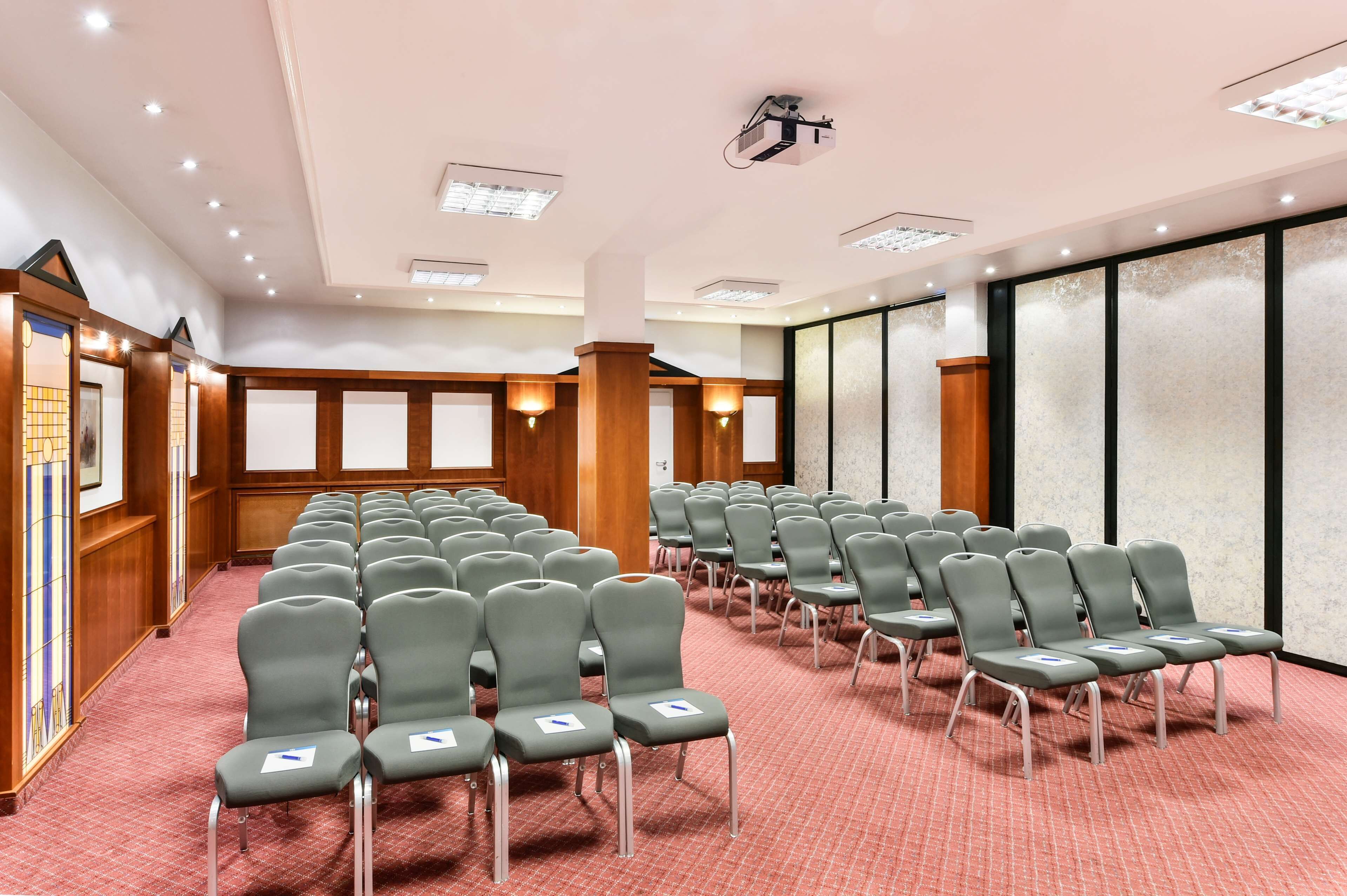 Une salle de conférence avec des rangées de chaises grises, un lambris en bois et un projecteur fixé au plafond.