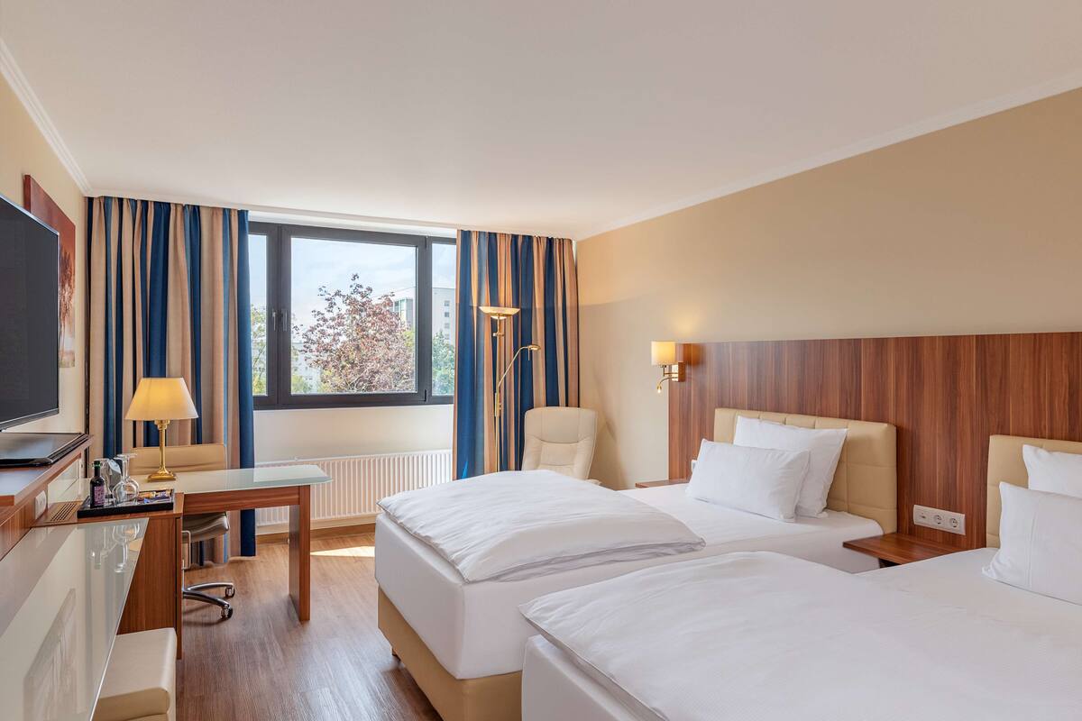 A hotel room with two beds, a desk, a chair, a TV, and a window with curtains.