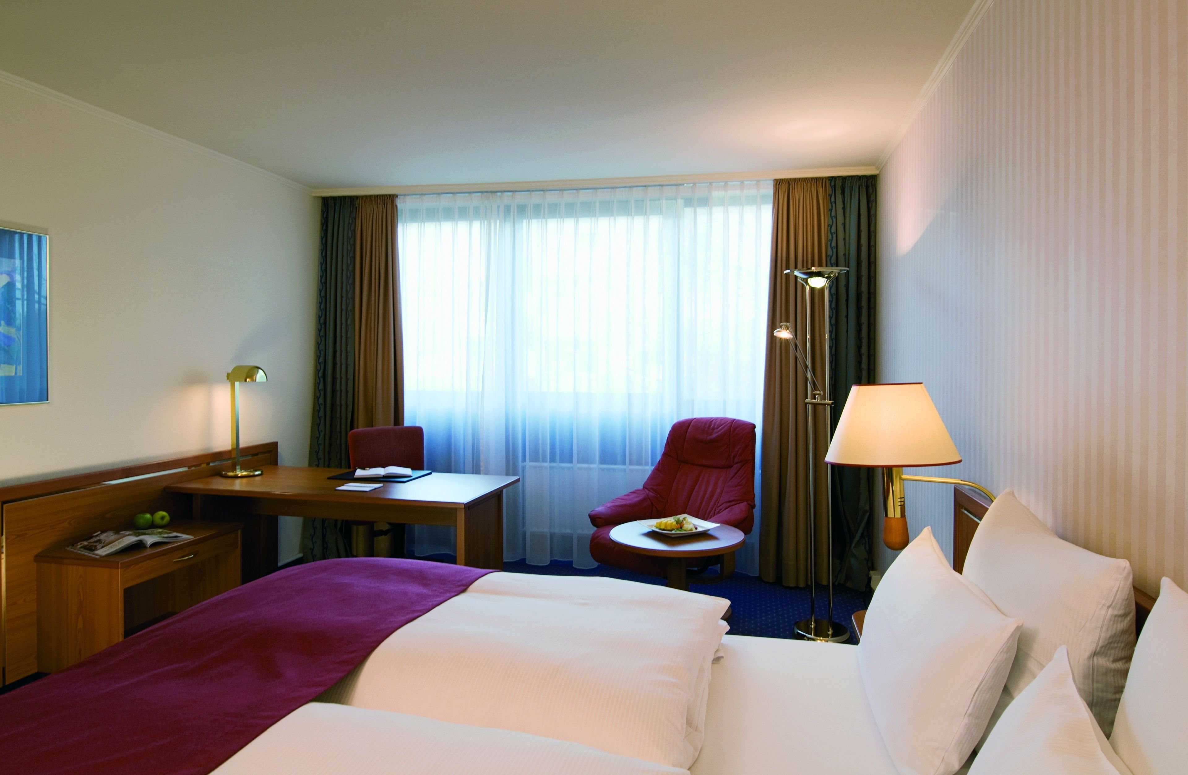 Een hotelkamer met een bed, een bureau, een stoel, een klein tafeltje met eten erop en een lamp.