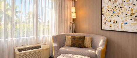 A hotel room with a large window, an air conditioning unit, a sofa with a cushion, a floor lamp, and a wall-mounted abstract painting.