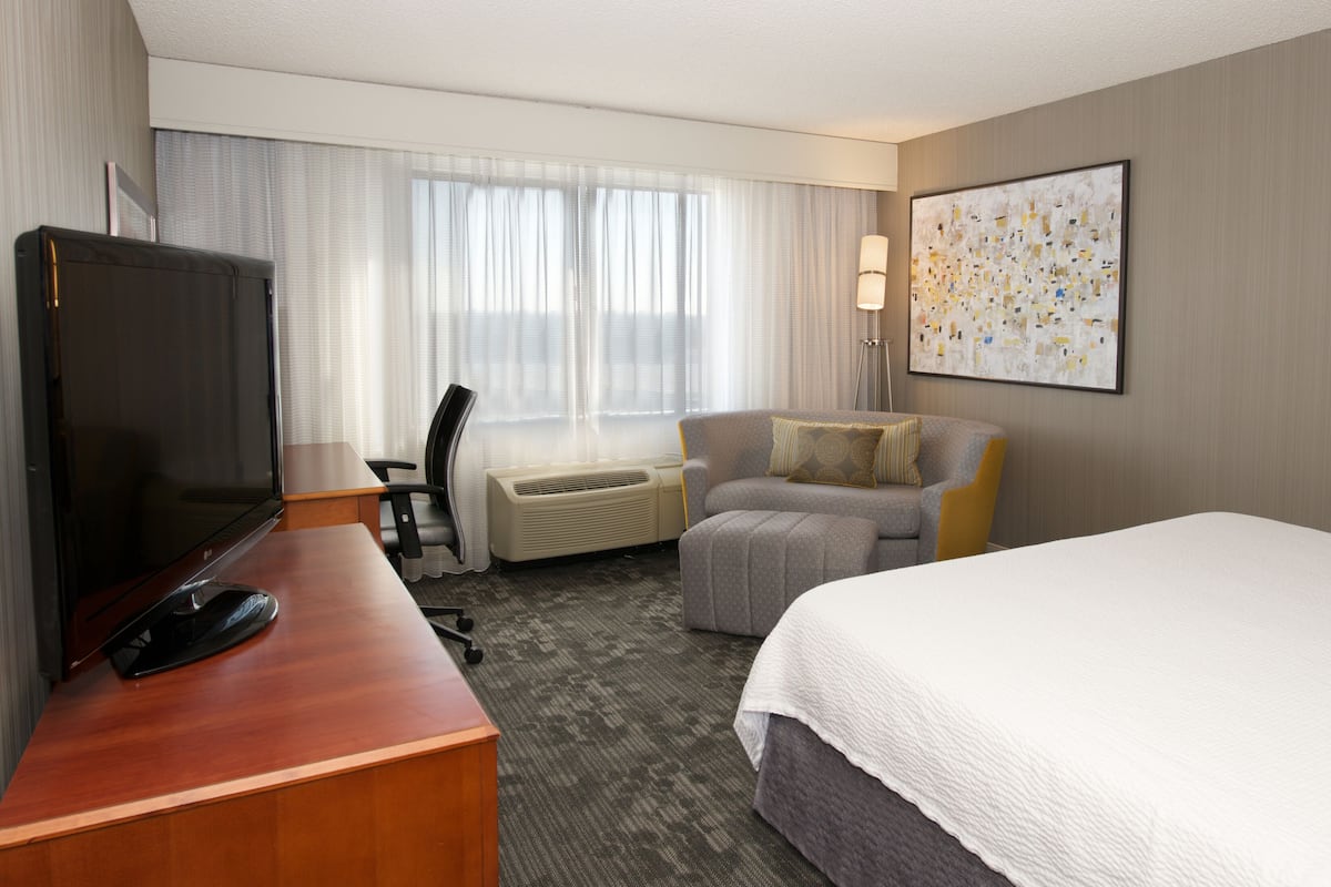 A hotel room with a bed, a desk with a computer, a chair, an air conditioner, and a large window with curtains.