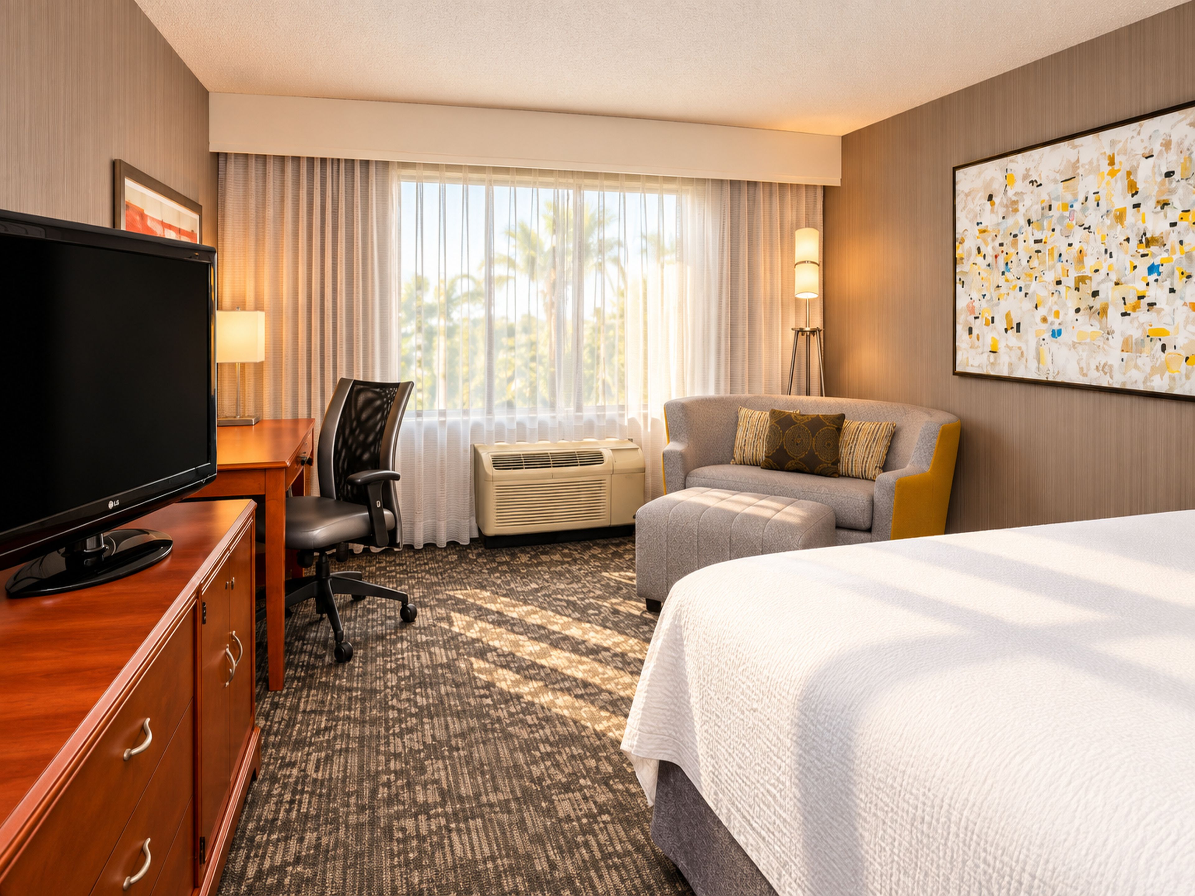 Ein Hotelzimmer mit einem Bett, einem Schreibtisch mit Fernseher, einem Sessel, einer Couch, einer Lampe und einem großen abstrakten Gemälde an der Wand.