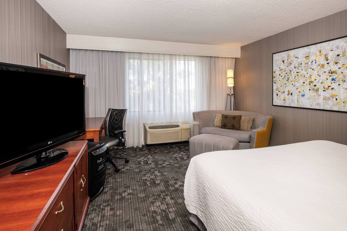 A hotel room with a bed, a desk with a computer, a TV, a chair, a lamp, and a large abstract painting on the wall.