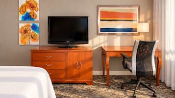 Quarto de hotel com cama, TV sobre um armário de madeira, escrivaninha com cadeira e obras de arte na parede.