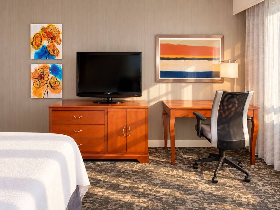 A hotel room with a bed, a TV on a wooden cabinet, a desk with a chair, and artwork on the wall.