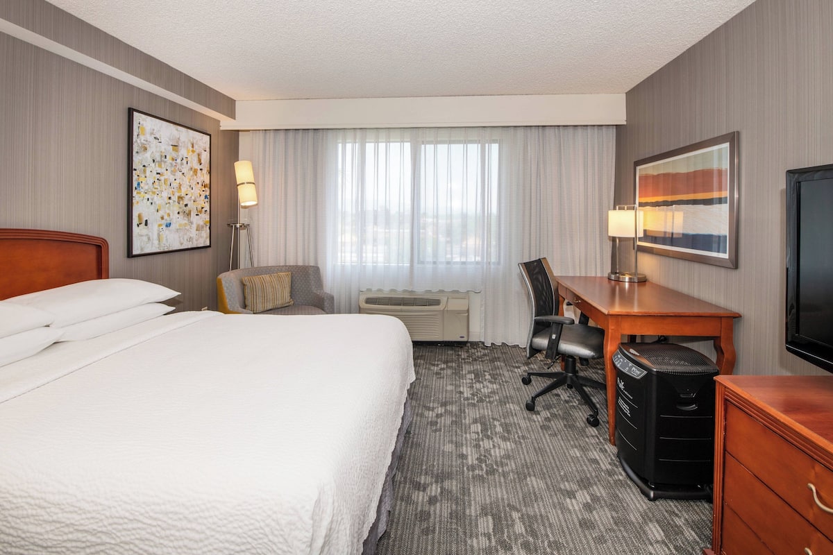A hotel room with a large bed, a desk with a computer, a chair, a TV, and a window with curtains.