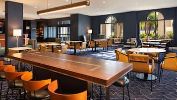 A modern hotel lobby with a central bar area, multiple seating arrangements, and large windows providing natural light.