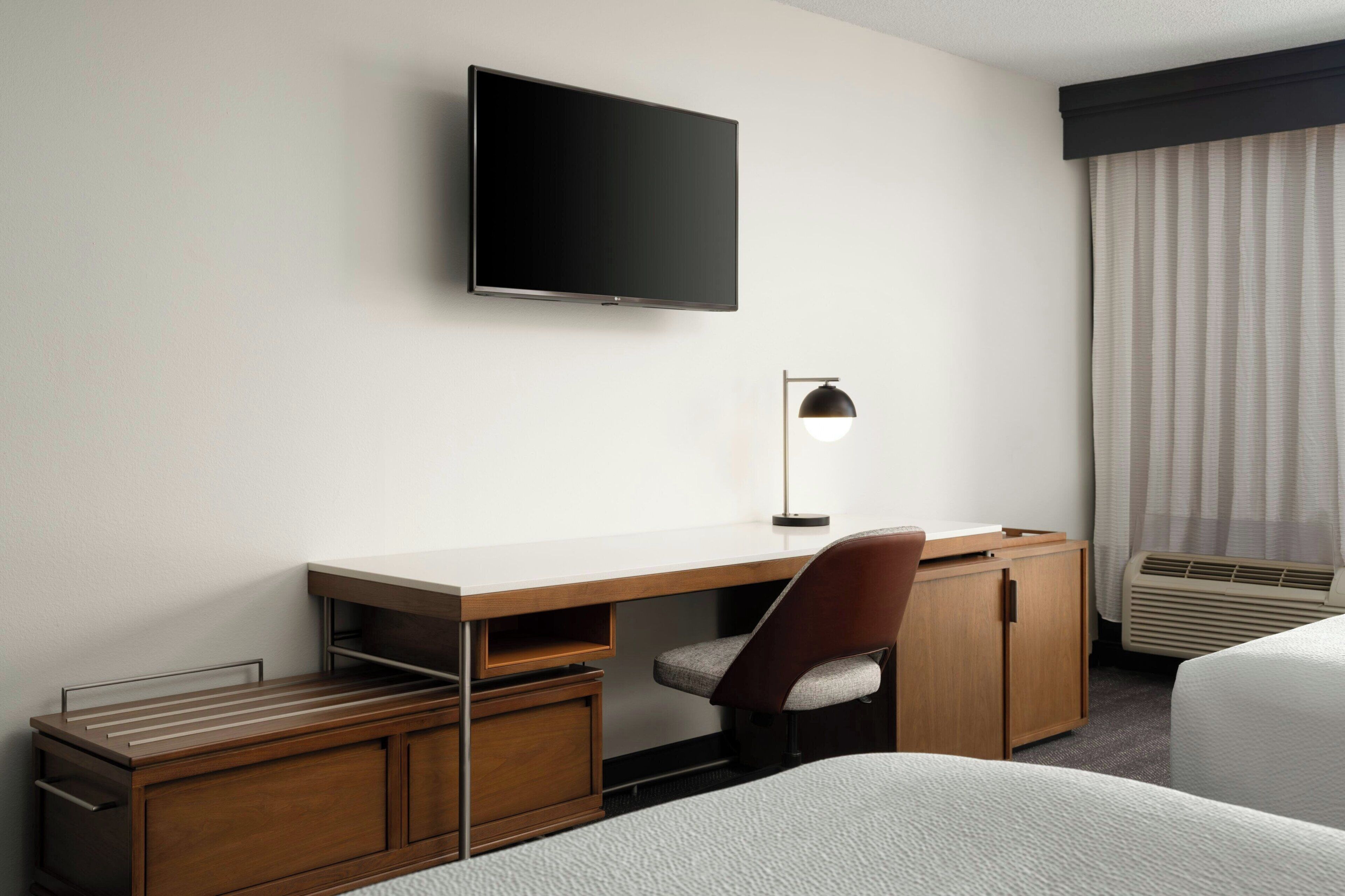 A hotel room with a flat-screen TV mounted on the wall, a desk with a chair, a bedside table, and a bed.