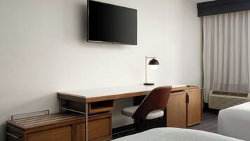 A hotel room with a flat-screen TV mounted on the wall, a desk with a chair, a bedside table, and a bed.
