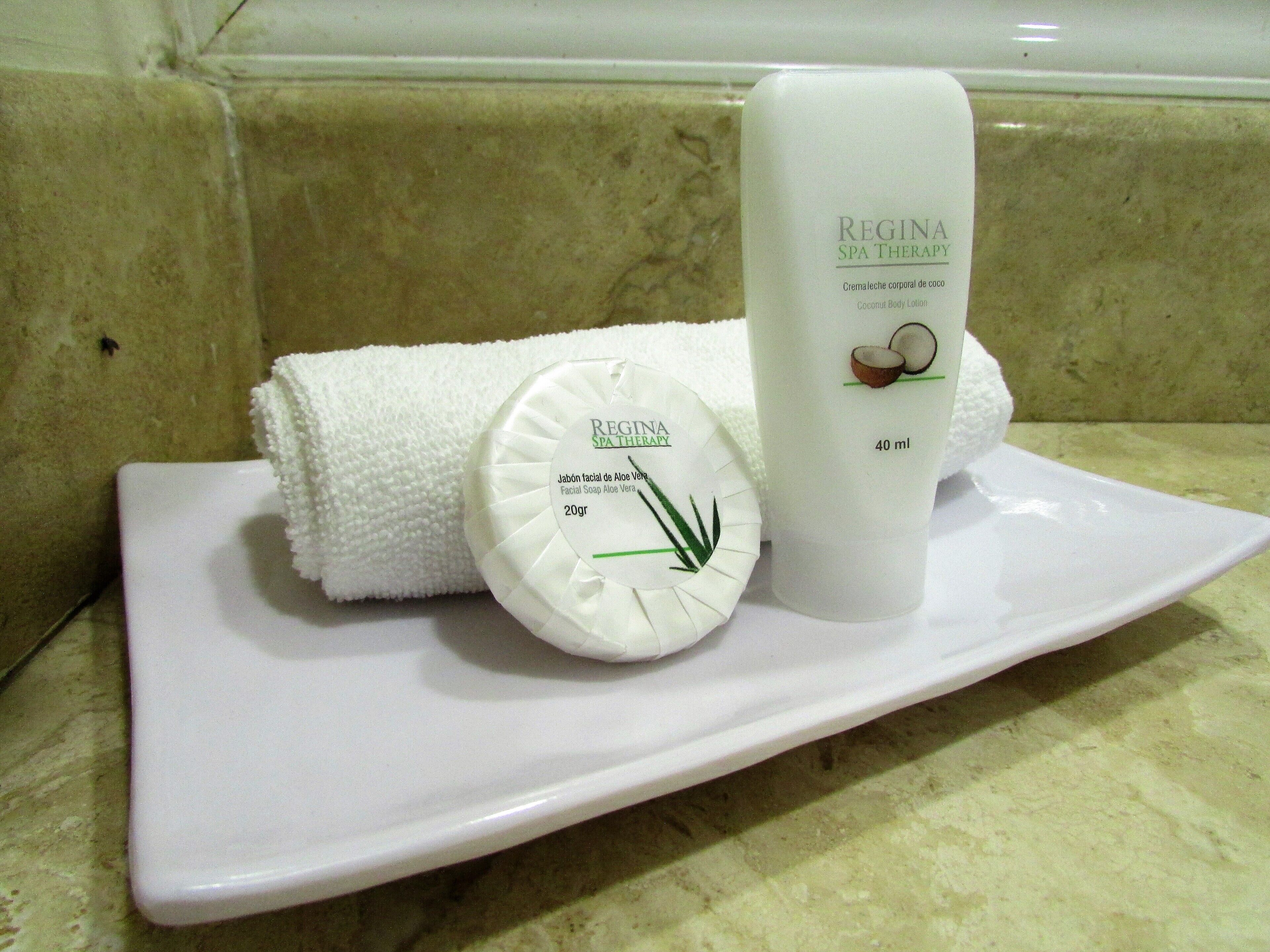 A white plate with a bottle of Regina Spa Therapy Coconut Body Lotion, a round soap bar, and a folded white towel.