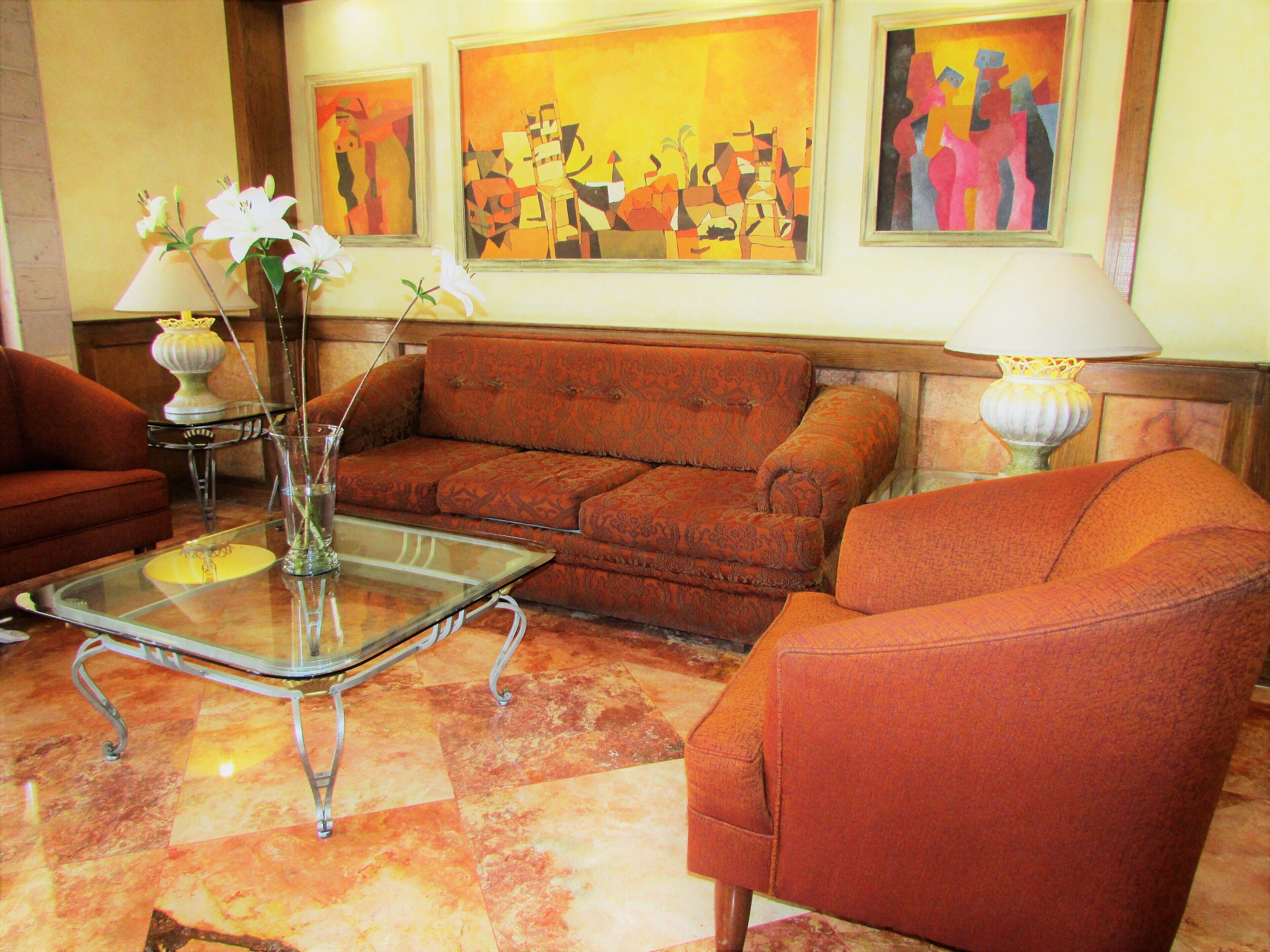 Una sala de estar con un sofá, sillones, una mesa de centro de vidrio y un florero con flores. Hay cuadros en la pared y una lámpara sobre una mesita auxiliar.
