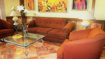 Una sala de estar con un sofá, sillones, una mesa de centro de vidrio y un florero con flores. Hay cuadros en la pared y una lámpara sobre una mesita auxiliar.