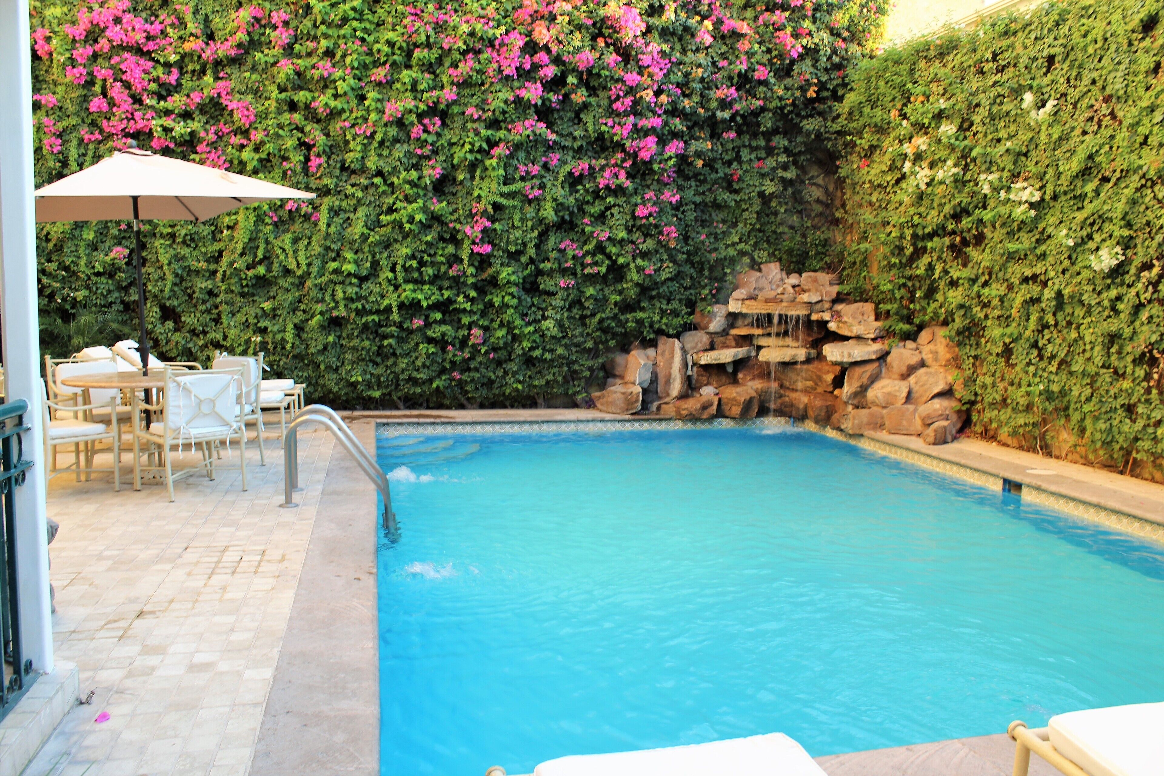 A swimming pool with a waterfall feature, surrounded by a lush green wall with pink flowers. There are lounge chairs and an umbrella on the patio area.