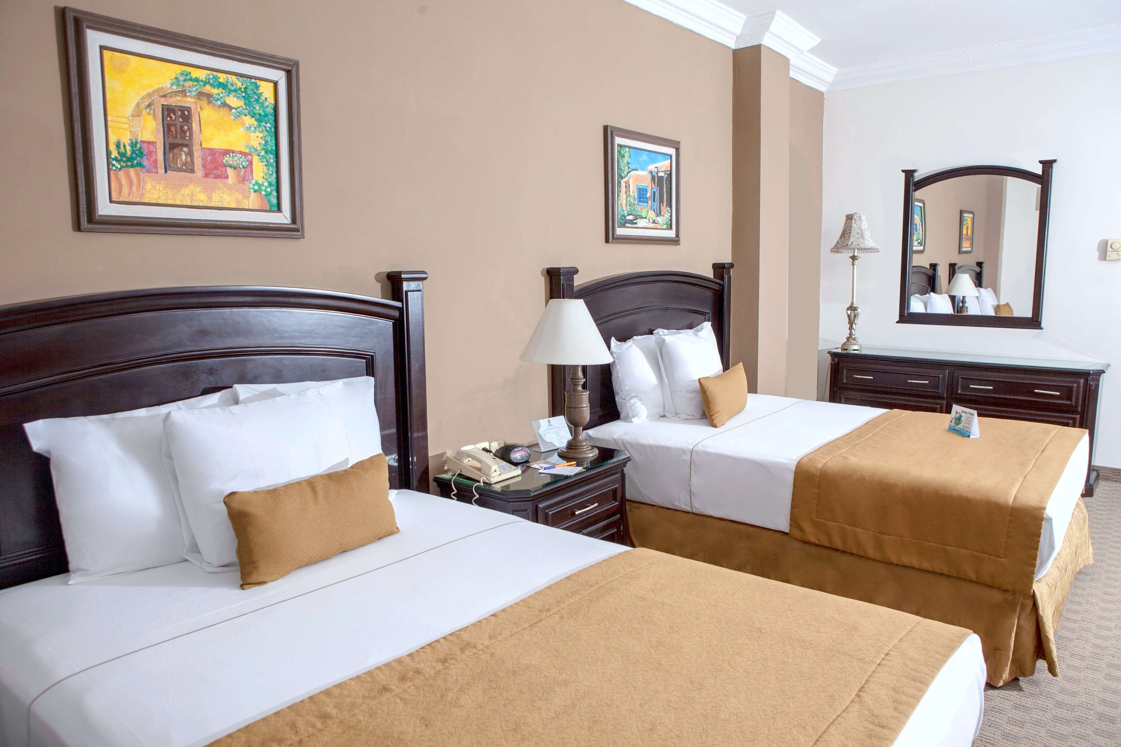A hotel room with two beds, a nightstand, a lamp, and framed artwork on the walls.