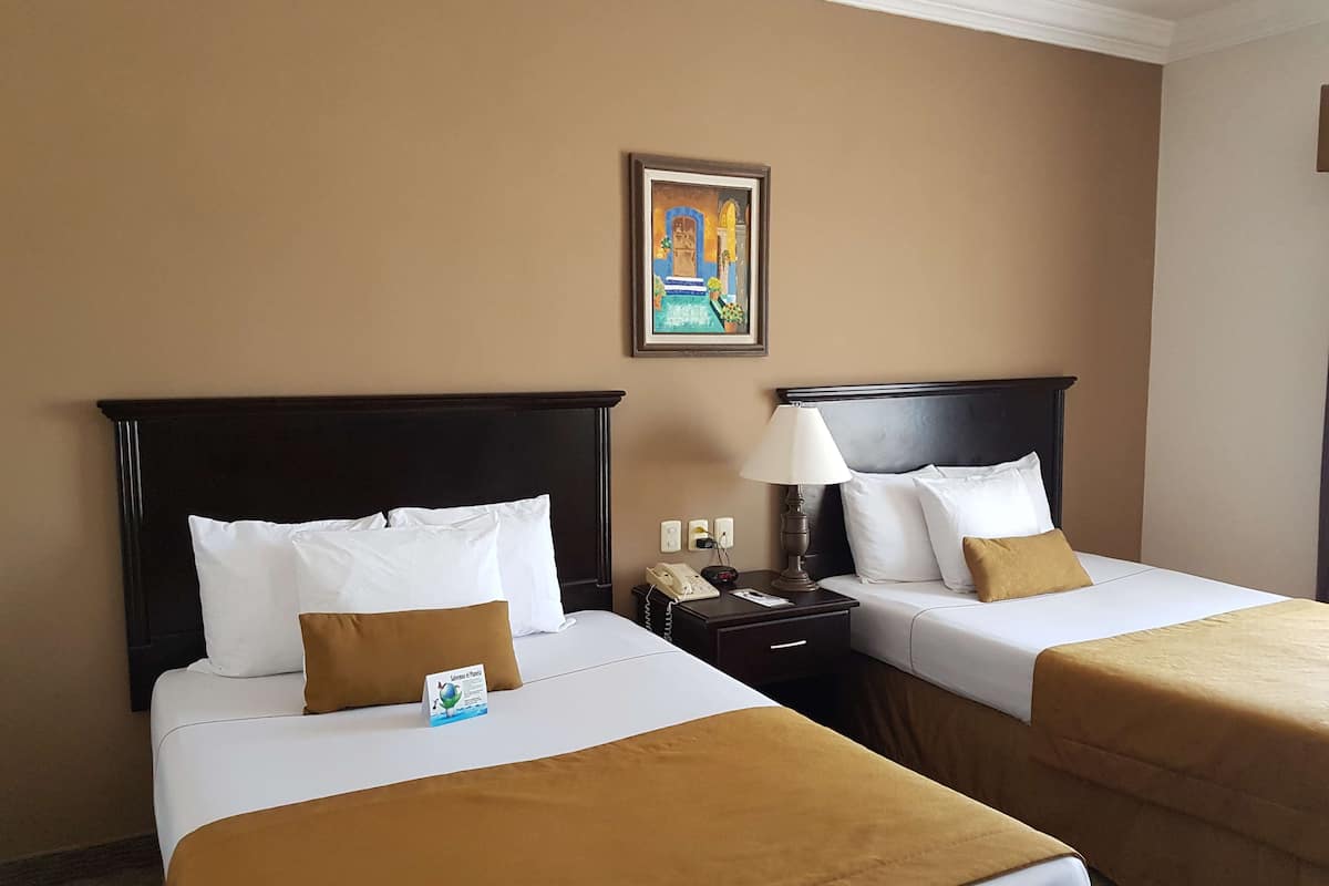 Une chambre d’hôtel avec deux lits, chacun doté de draps blancs et d’une tête de lit marron. Une table de chevet avec une lampe et un téléphone se trouve d’un côté, et une image encadrée est accrochée au mur au-dessus des lits.