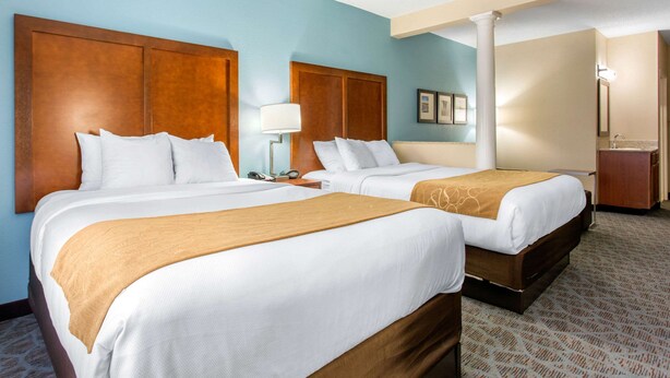 A hotel room with two beds, a nightstand, a lamp, and a bathroom with a mirror and sink. - Comfort Suites Southport - Oak Island (Southport)