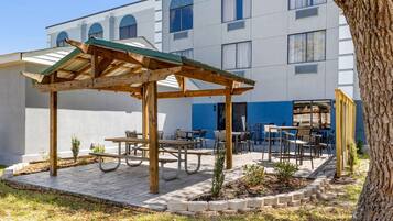 A covered outdoor seating area with picnic tables and chairs, surrounded by a low stone wall and a tree.