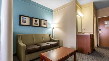A hotel room with a sofa, a wooden table, a lamp, a mirror, and a small bathroom area.