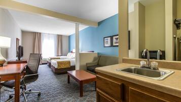 Quarto de hotel com cama, escrivaninha, cadeira e cozinha compacta.