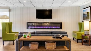 A modern living room with a flat-screen TV mounted on the wall, a built-in fireplace with a purple light, a sofa, two green armchairs, and a wooden coffee table with baskets. - Comfort Suites Southport - Oak Island (Southport)