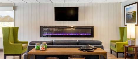 A modern living room with a flat-screen TV mounted on the wall, a built-in fireplace with a purple light, a sofa, two green armchairs, and a wooden coffee table with baskets.