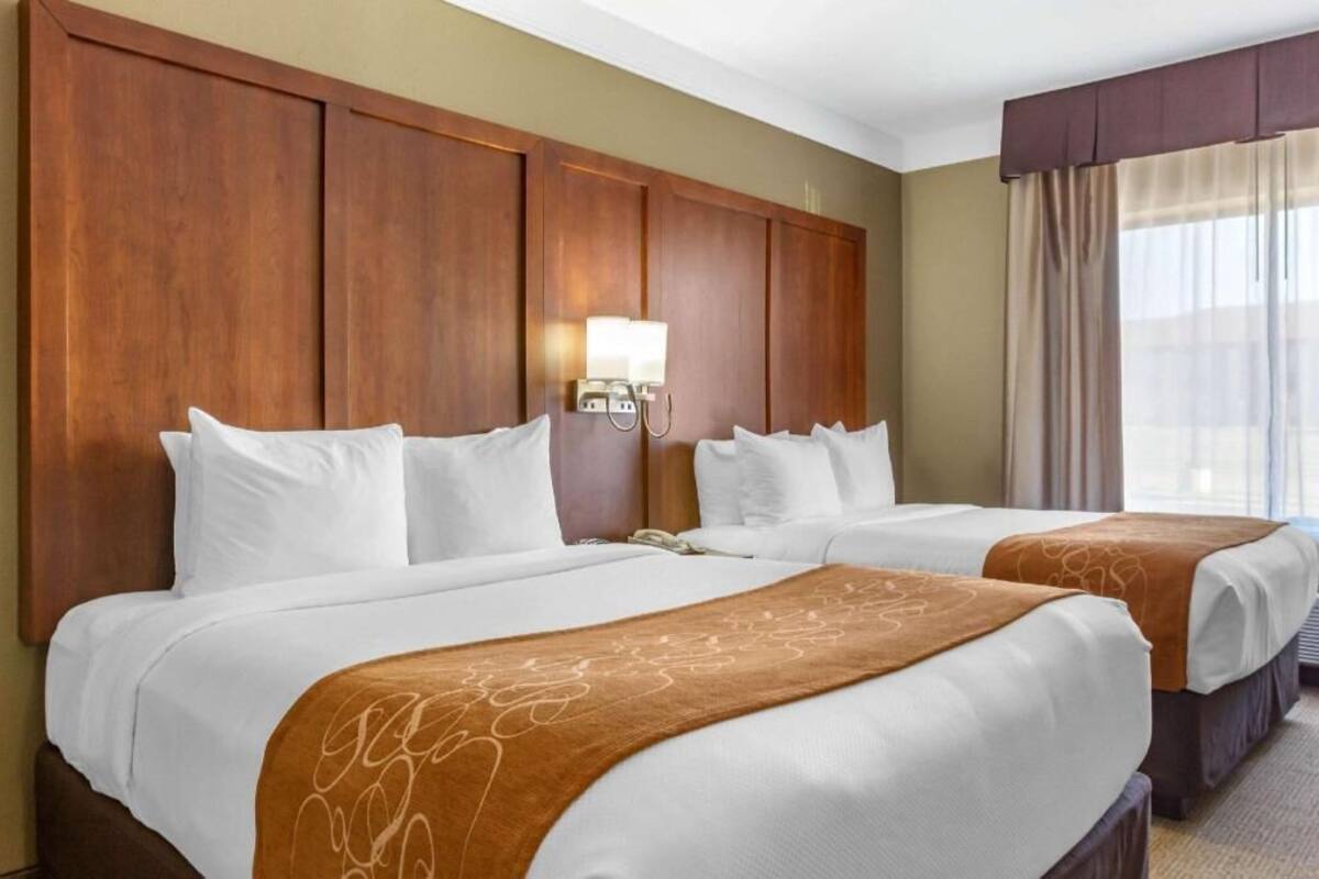 Habitación de hotel con dos camas, cabecera de madera, mesita de noche, lámpara y ventana con cortinas.