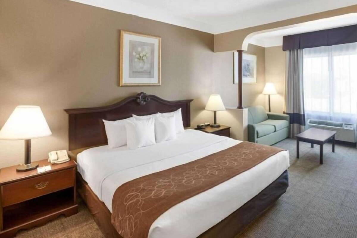 Una habitación de hotel con cama, lámparas de noche, escritorio, silla y un amplio ventanal con cortinas.