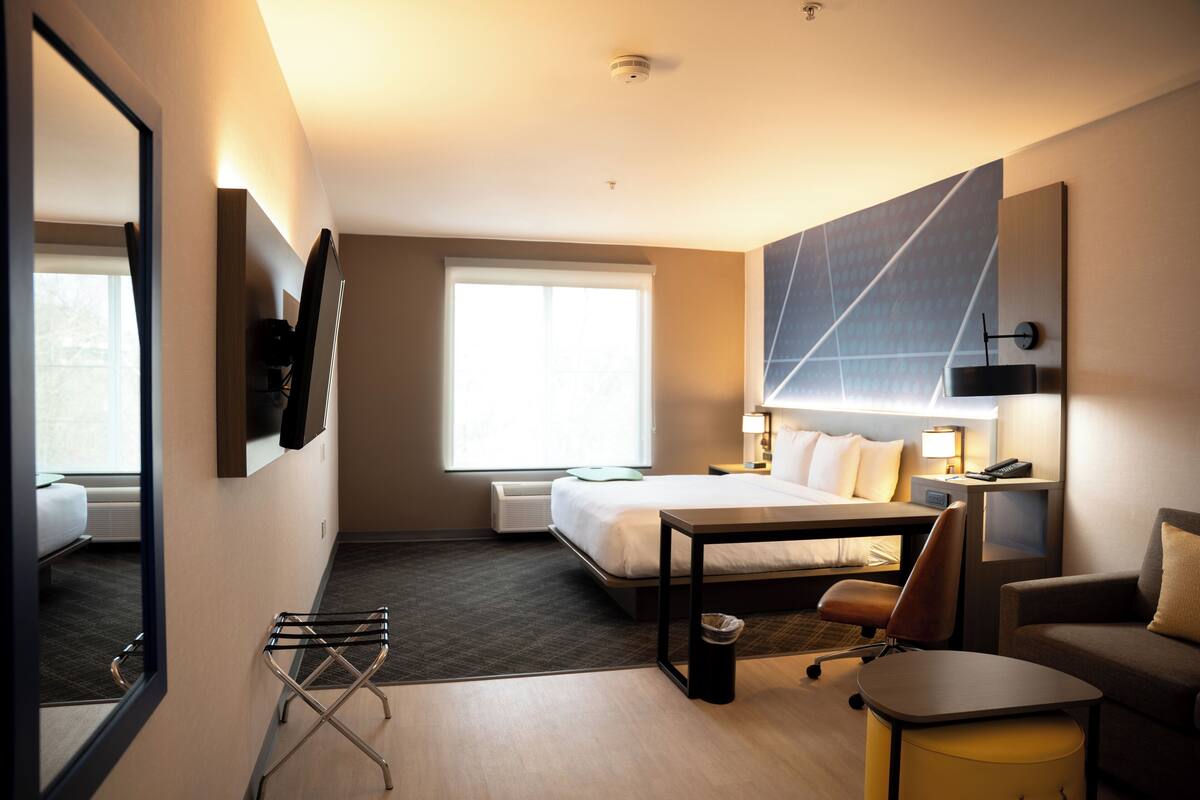 Hotellihuone, jossa on sänky, työpöytä, tuoli, pieni pöytä, lamppu, televisio ja ikkuna.