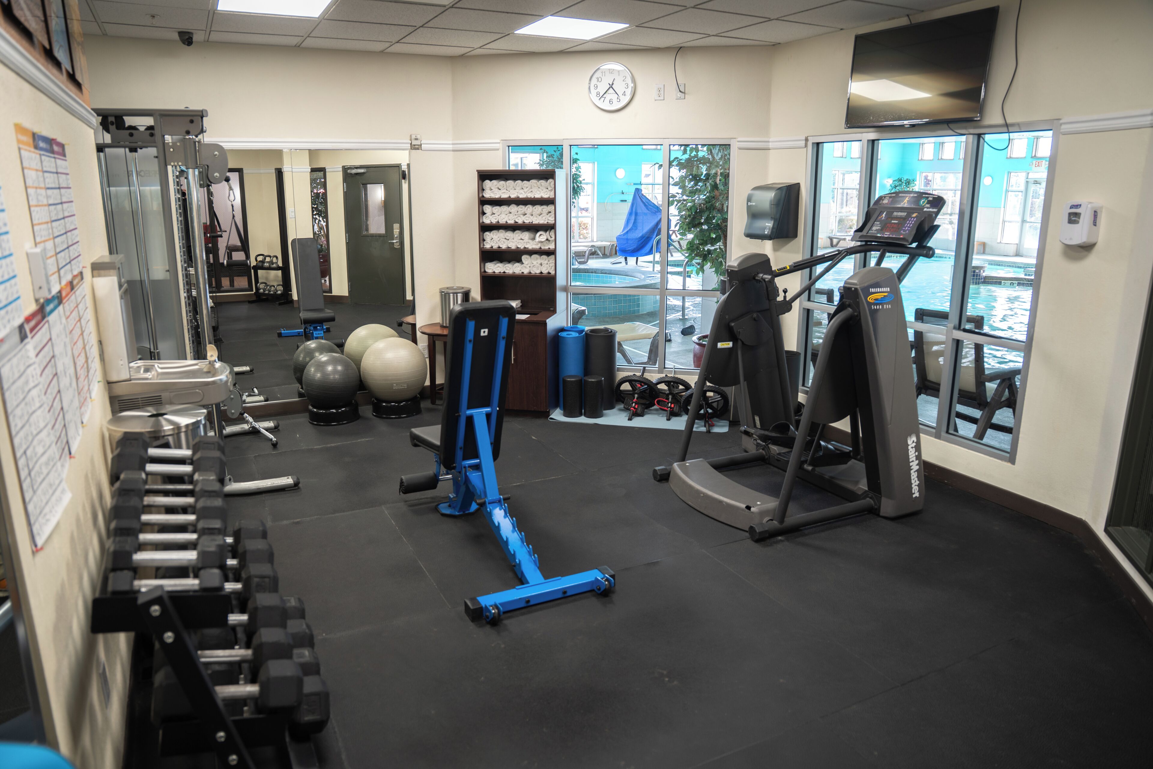 A gym with exercise equipment, a clock, and a window with a view of a pool.