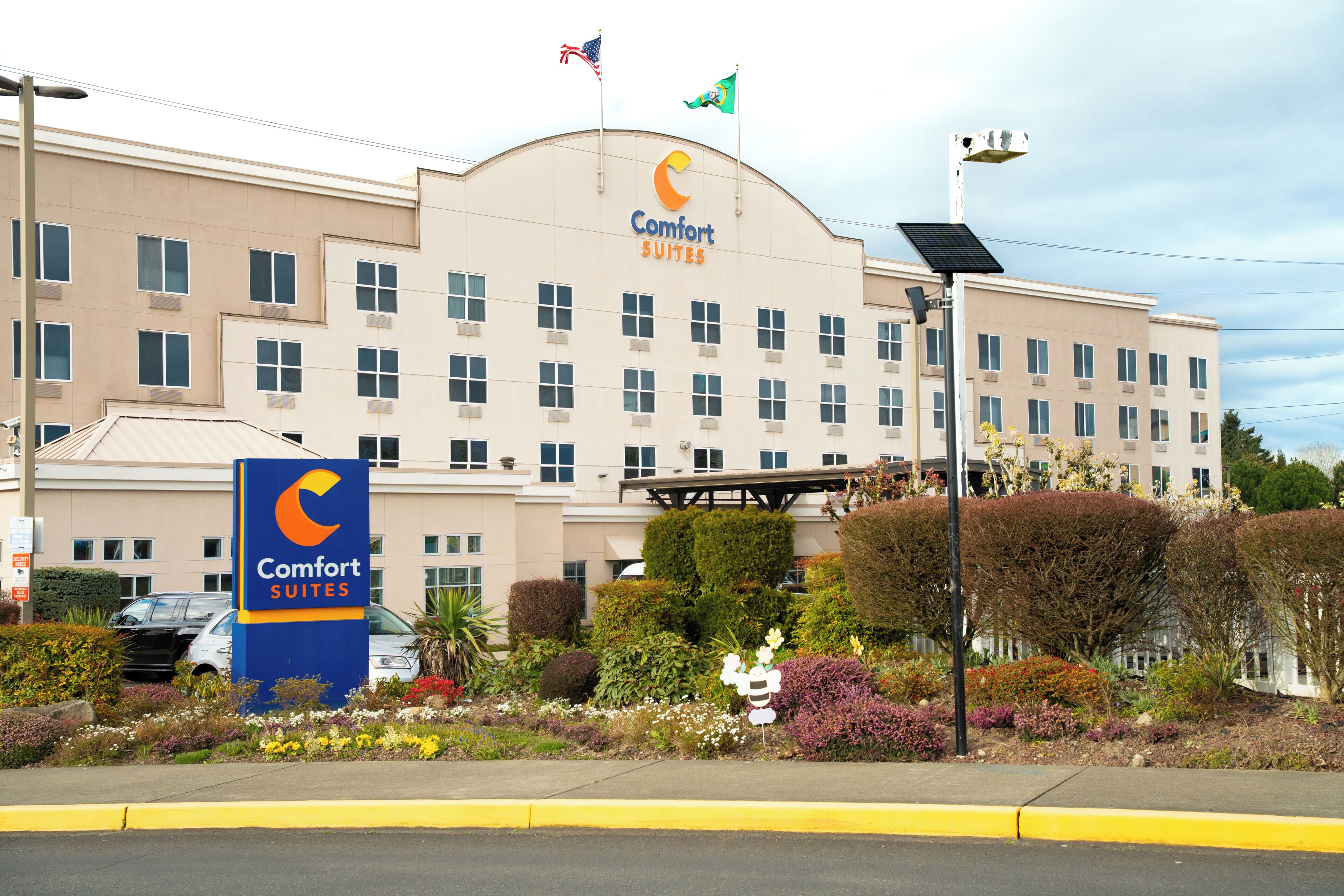 Un hotel con un letrero grande que dice 'Comfort Suites', varias banderas y un jardín bien cuidado.