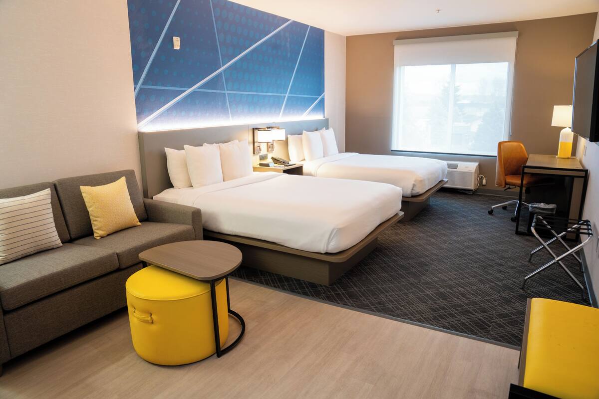 Hotellihuone, jossa on kaksi sänkyä, sohva, pieni pöytä, tuoli ja työpöytä, jossa on lamppu.