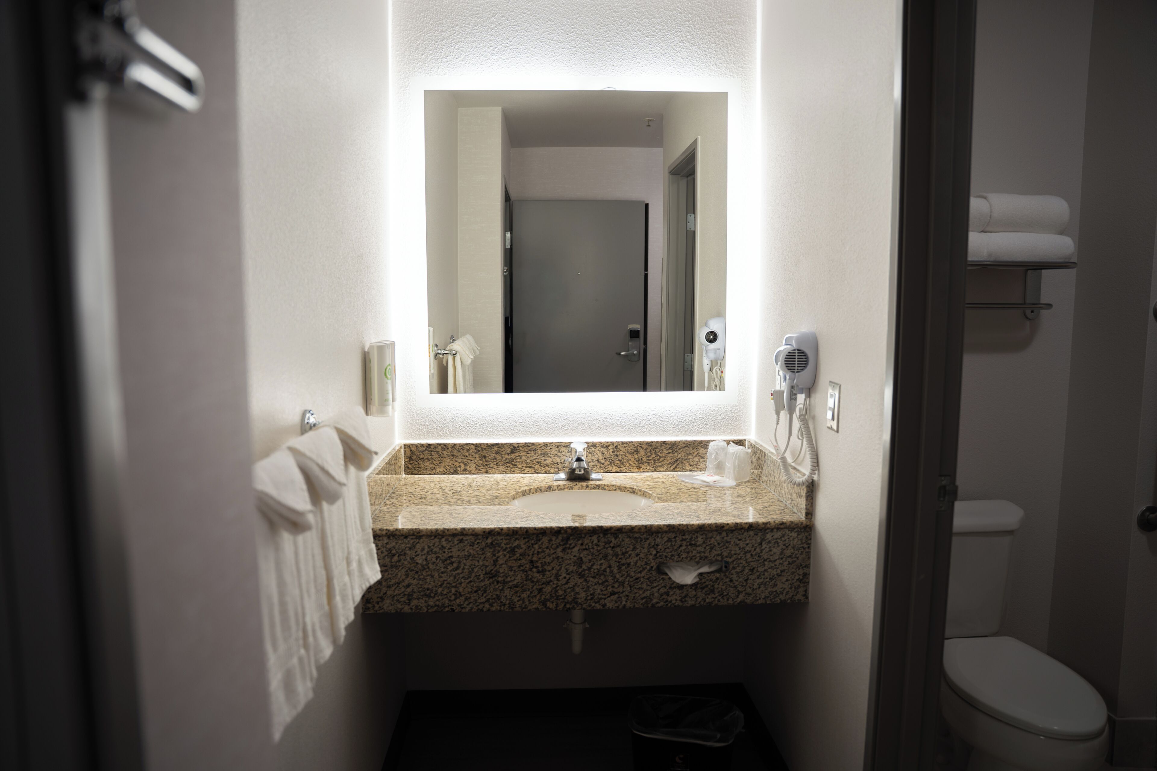 A bathroom with a granite countertop, a mirror, a towel rack, a hairdryer, and a toilet.