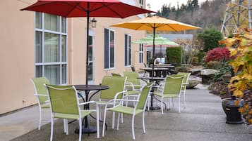 Un patio al aire libre con mesas y sillas, dos sombrillas y un jardín con follaje otoñal.