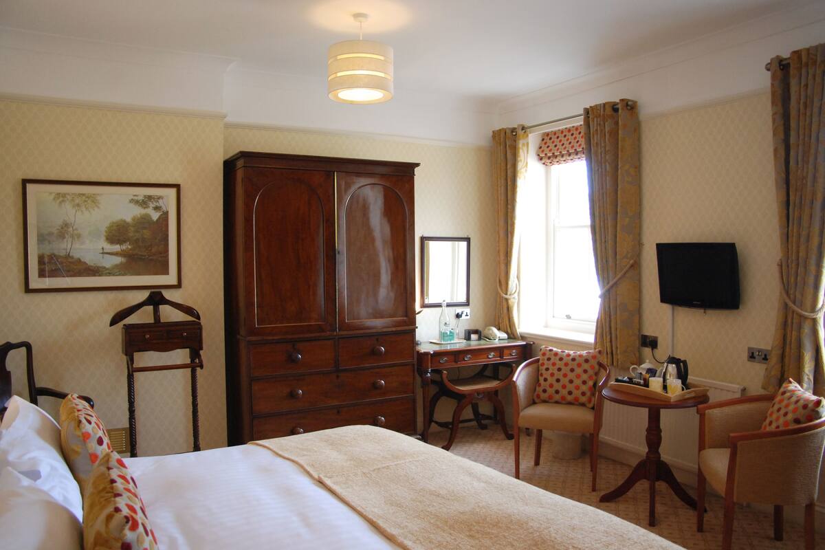 A hotel room with a large bed, a wooden wardrobe, a desk with a chair, a television, and a window with curtains.
