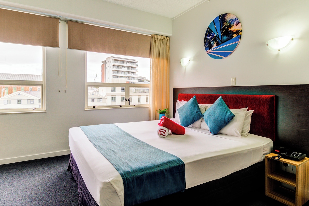A hotel room with a large bed, a view of buildings through the window, a clock on the nightstand, and a wall-mounted light fixture.