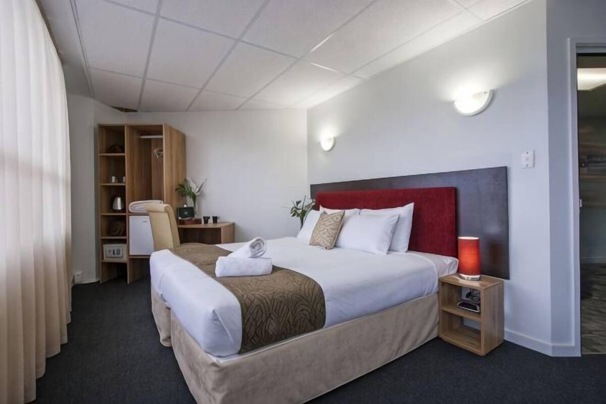 A hotel room with a large bed, a desk with a chair, a wardrobe, and a bedside table with a lamp.