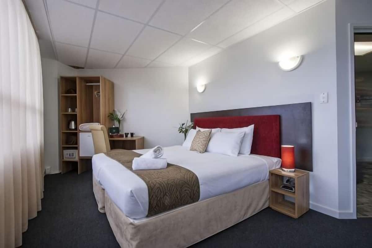 A hotel room with a large bed, a desk with a chair, a wardrobe, and a bedside table with a lamp.