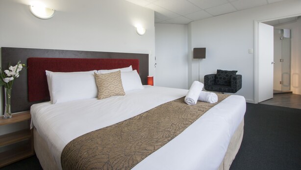 Una habitación de hotel con una cama grande, un cabecero rojo, una mesita de noche con un jarrón de flores y una silla con un cojín estampado.