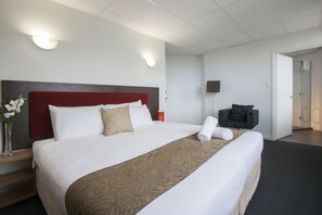 A hotel room with a large bed, a red headboard, a bedside table with a vase of flowers, and a chair with a patterned cushion.