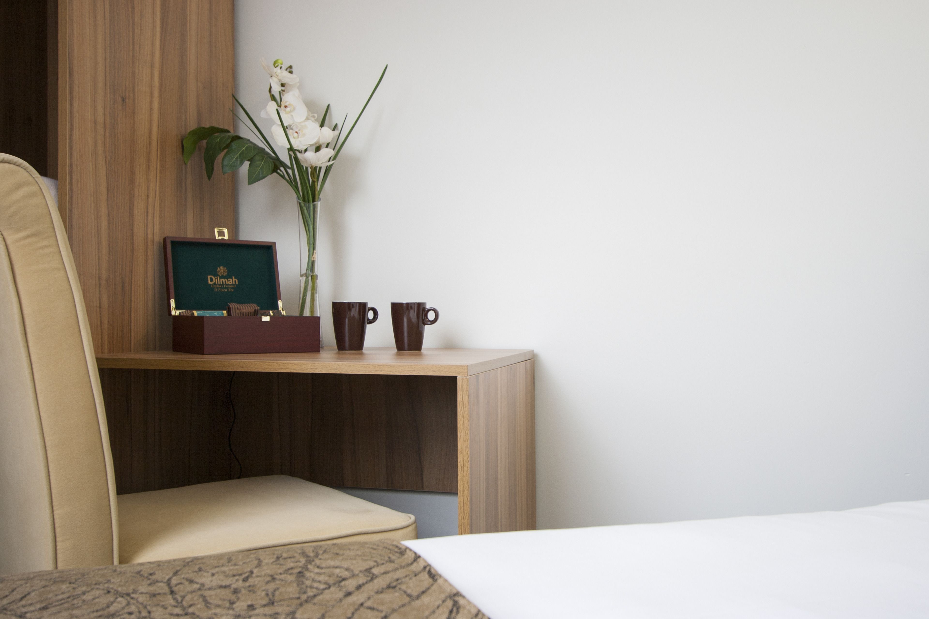 Habitación de hotel ordenada con un escritorio de madera, un certificado enmarcado, dos tazas y un jarrón con flores blancas.